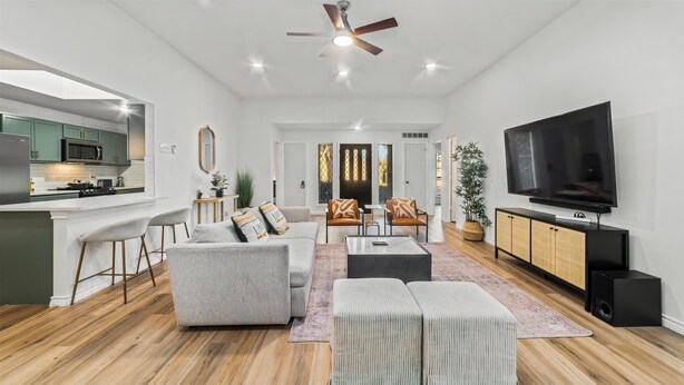 A modern living room with a grey sofa, a coffee table, and a TV. There is a kitchen area with a microwave and a bar counter. - Exquisite 4 Bed Great Location (Richardson)