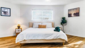 A neatly arranged bedroom with a bed, a nightstand with a lamp, a window with blinds, and two framed artworks on the walls. - Exquisite 4 Bed Great Location (Richardson)