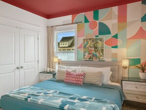 Un dormitorio con cama, mesita de noche, lámpara, ventana con vistas a una casa y un cuadro abstracto y colorido en la pared.