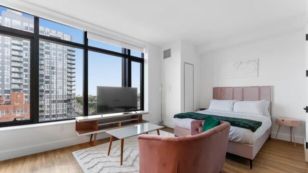 A modern bedroom with a large window, a bed, a TV on a stand, and a pink chair. - Chic studio with fitness room, WiFi in delightful Brooklyn (Brooklyn)
