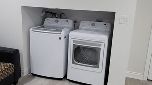 A laundry room with two white front-loading washing machines and a dryer, wooden flooring, and a black couch with a patterned cover. - Cozy Private Oasis Peaceful Stay (Milton)