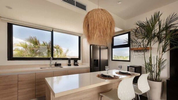 A modern kitchen with a large island, a hanging light fixture, and a view of palm trees through large windows. - Aura Luna Tropical Rooftop Beach Club Access (Tulum)