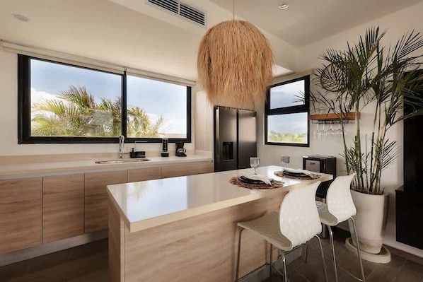 A modern kitchen with a large island, a hanging light fixture, and a view of palm trees through large windows.