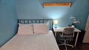 A neatly made bed with white linens and pillows, a desk with a chair, a lamp, and a small potted plant.