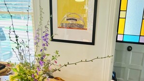 Uma mesa de madeira redonda com um vaso de flores branco, duas cadeiras de madeira e um quadro pendurado na parede.