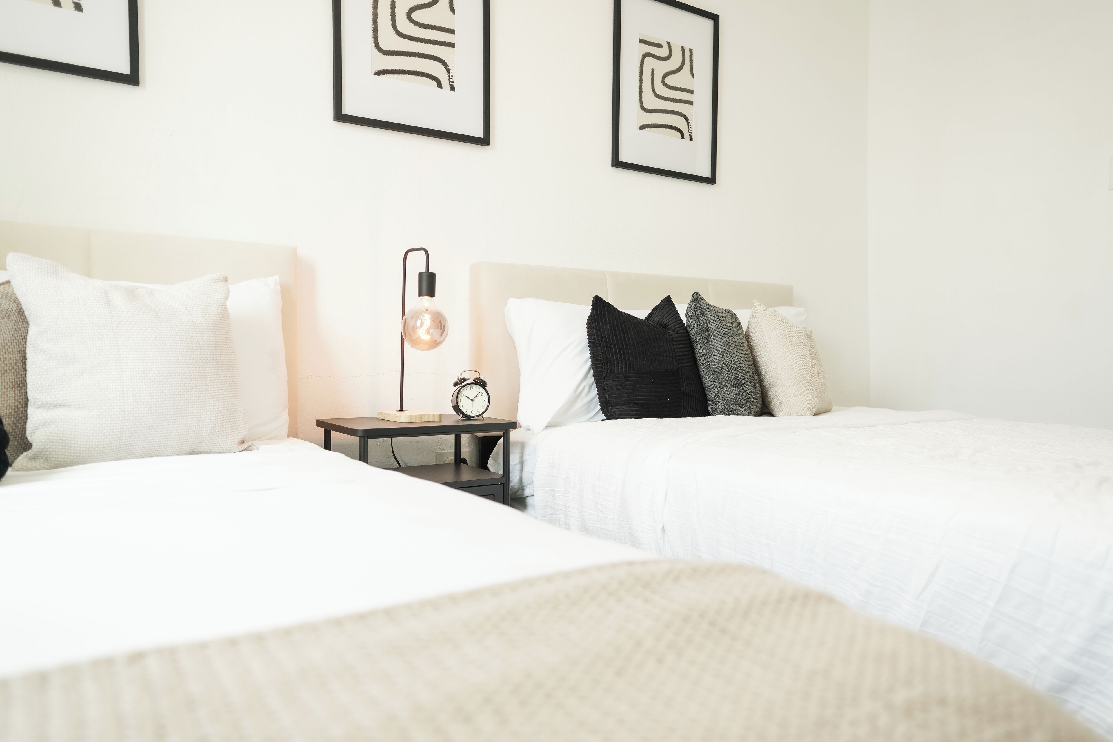 A hotel room with two beds, a bedside table with a lamp, and framed artwork on the wall.