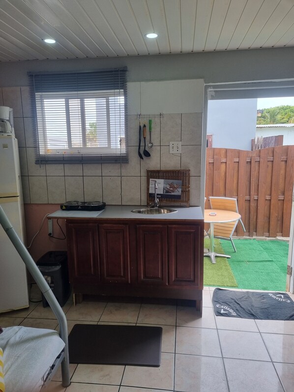 A compact kitchen with a built-in sink, a window with blinds, and a small dining area with a chair.