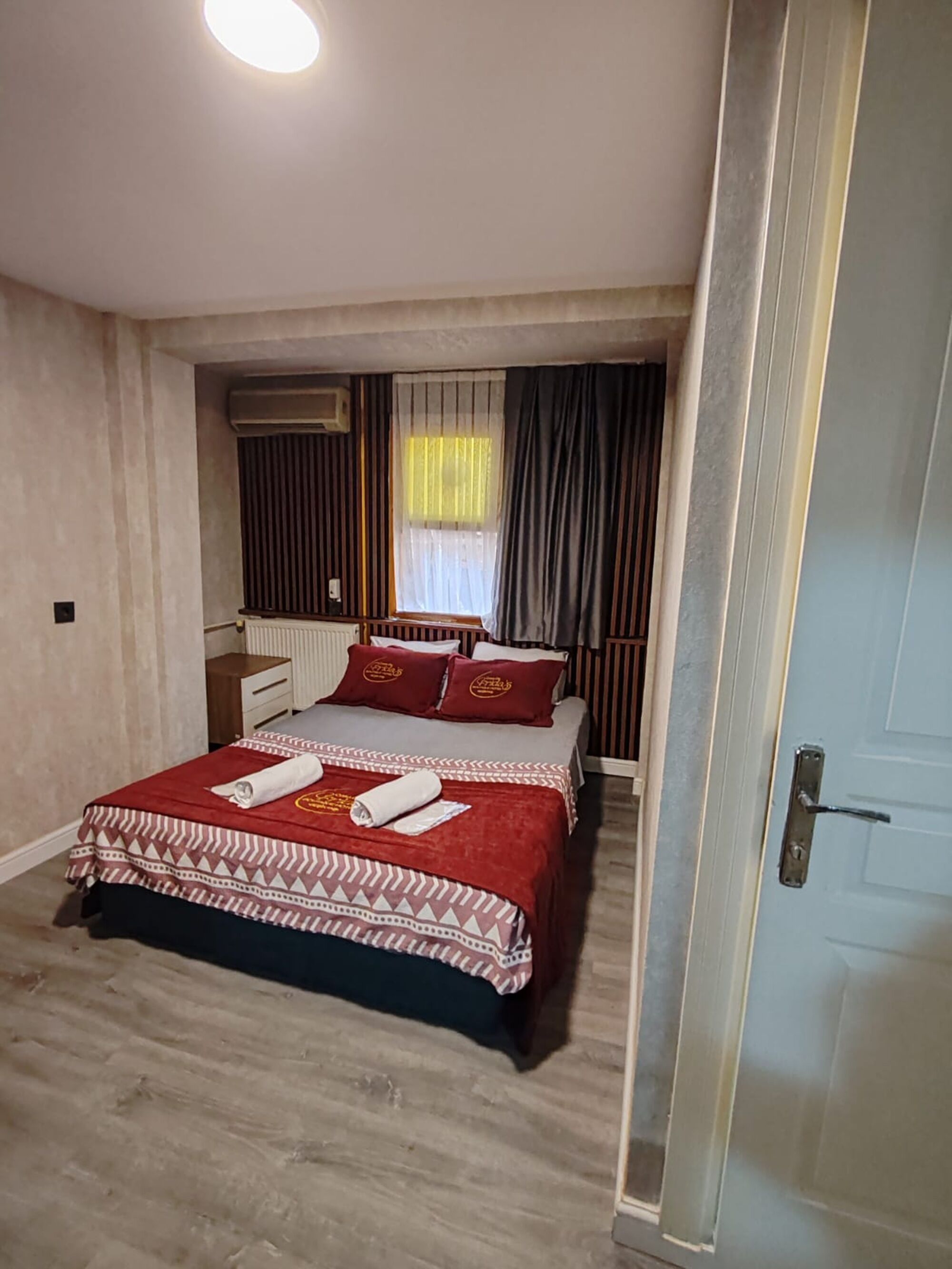 A hotel room with a bed, a bedside table, a window with curtains, and a door.