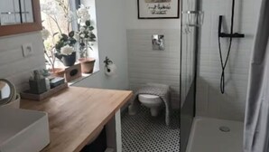Un baño compacto con tocador de madera, inodoro pequeño, ducha y ventana con plantas en macetas.