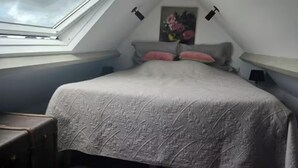 Un dormitorio con tragaluz, una cama con colcha estampada, un cuadro en la pared y una mesita con lámpara.