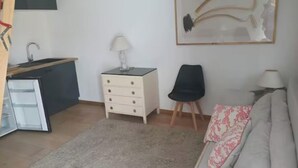 Un espacio habitable compacto con kitchenette, un sofá con cojines estampados, una pequeña mesita auxiliar y una silla.