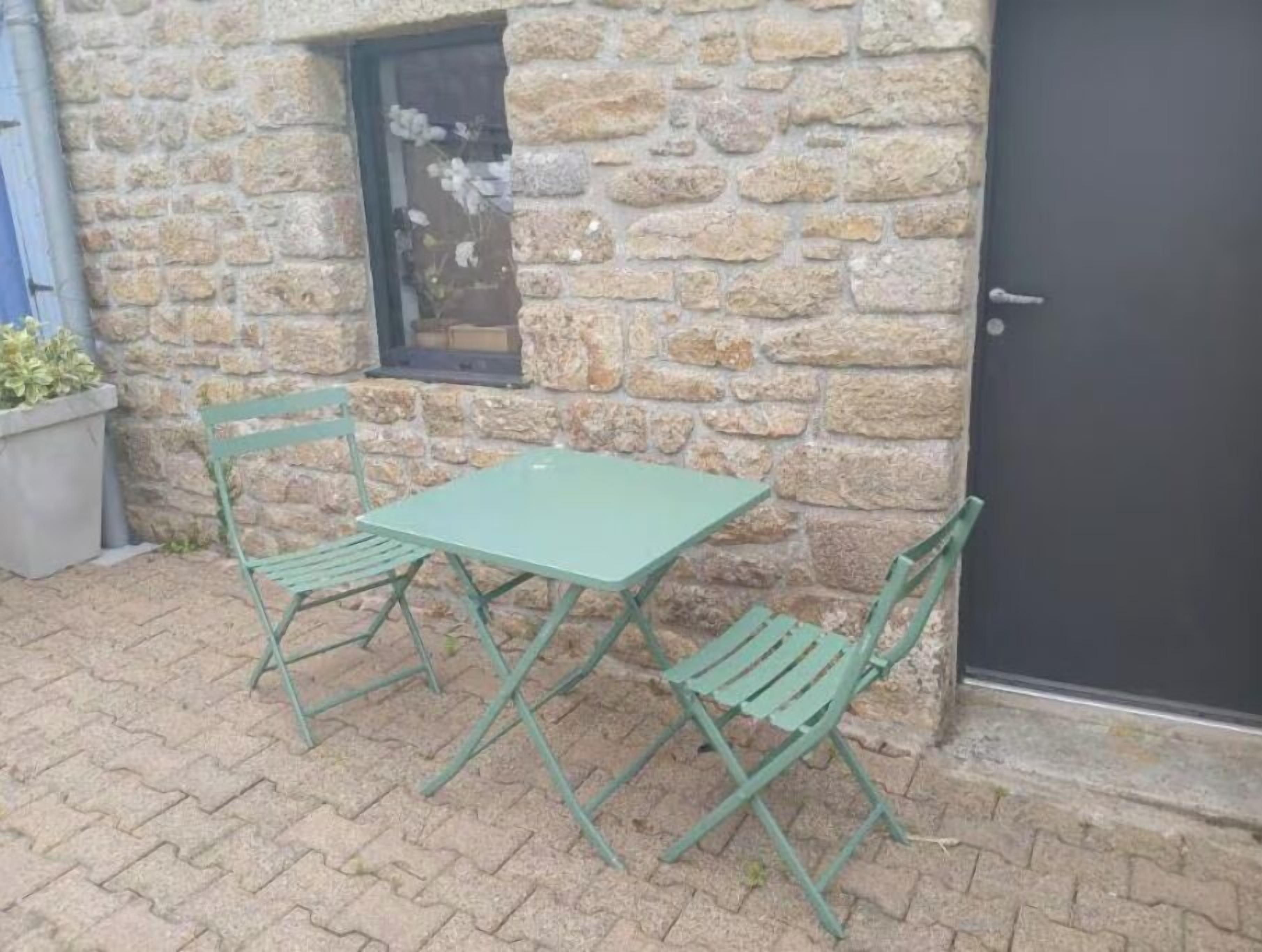 A stone building with a small window, a door, and a green table and chairs set on a paved patio.