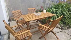 Un conjunto de mesa y sillas de madera frente a un muro de hormigón, con un pequeño jardín con flores y plantas.