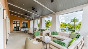 Un espacio lounge al aire libre con techo, con asientos y mesas, ventanas amplias y vistas a palmeras.