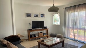 En stue med sofa, sofabord, fjernsyn på træstel og et vindue med lette gardiner.