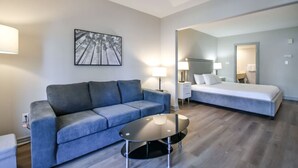 Une chambre d’hôtel moderne avec un canapé gris, une table ronde, un lit et un grand miroir.