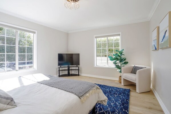 A modern bedroom with a bed, a TV on a stand, a sofa, and large windows.