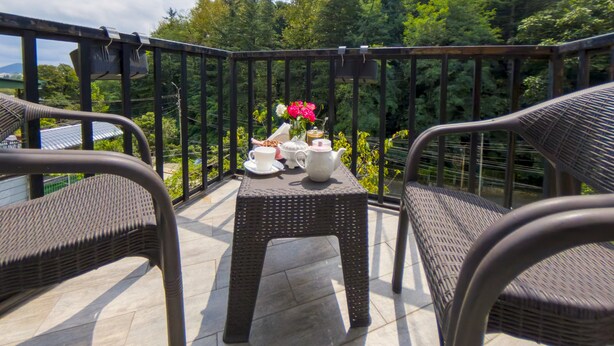 Un balcón con mobiliario de mimbre, una mesa con tetera, tazas y flores, y vista a un área boscosa.