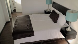 A neatly made bed with a dark brown bedspread and white linens, two bedside tables with lamps, and a door leading to another room.