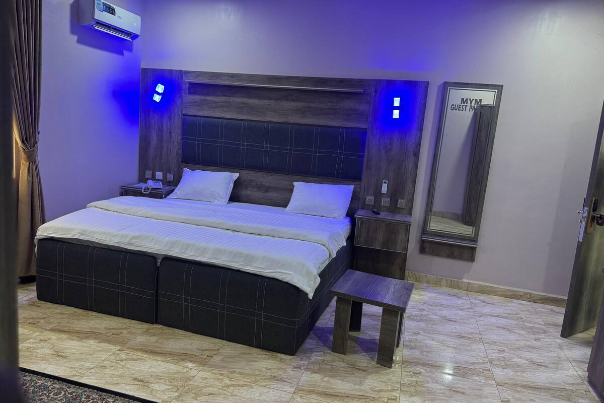 Un dormitorio moderno con una cama grande, mesitas de noche, un pequeño banco, un equipo de aire acondicionado y un espejo.