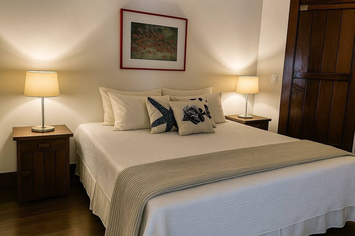 Een net opgemaakt bed met een beige dekbed en kussens, twee nachtkastjes met lampen, een lijst met een kunstwerk aan de muur, en een houten deur.