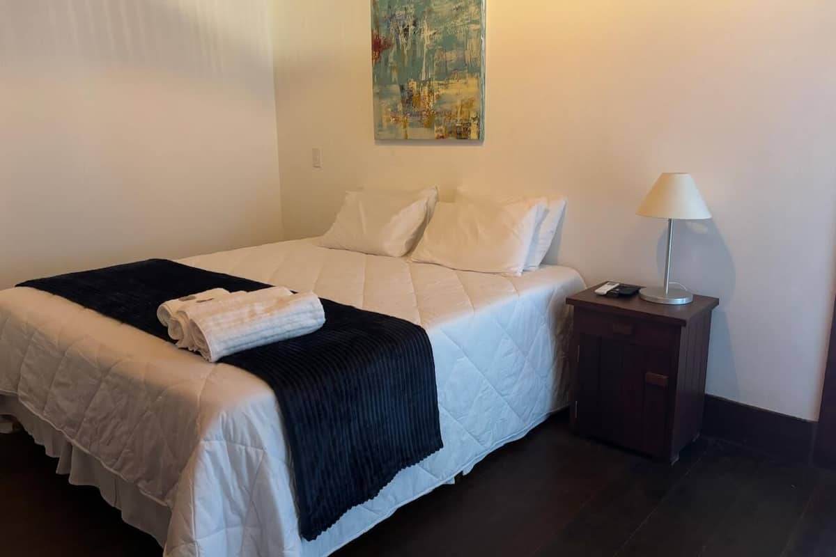 Een net opgemaakt bed met witte lakens en een donkere plaid, een bedlampje en een klein houten nachtkastje.