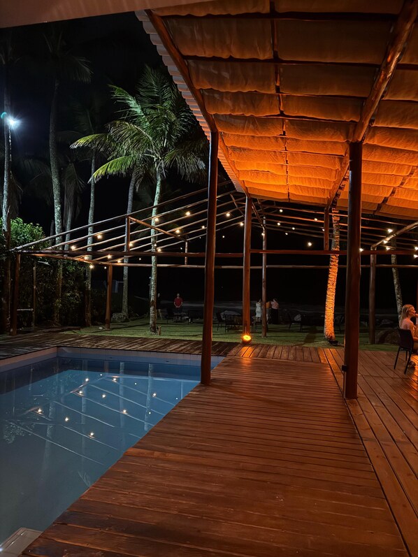 An outdoor pool area with a wooden deck, a covered seating area with string lights, and palm trees in the background. - 120 Hotel & Beach Club (Guaruja)