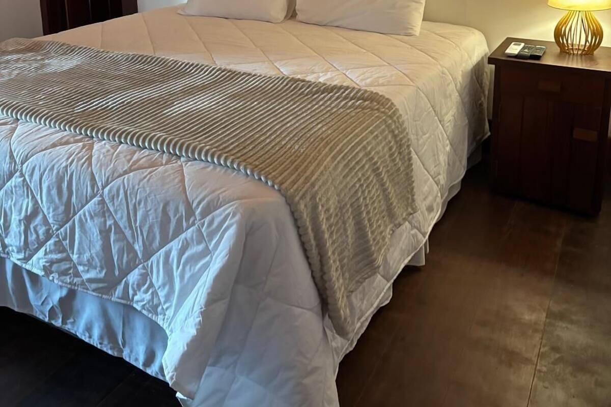 Een net opgemaakt bed met een gewatteerde witte dekbedovertrek, een houten nachtkastje met een lamp, en een ingelijst kunstwerk aan de muur.