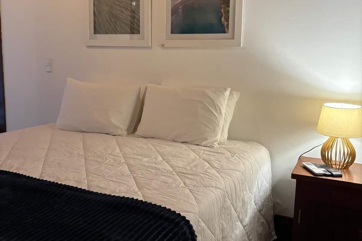 Een netjes opgemaakt bed met een quilt, twee kussens, een zwarte plaid en een nachtkastje met een lamp. Boven het bed hangen twee ingelijste foto’s aan de muur.