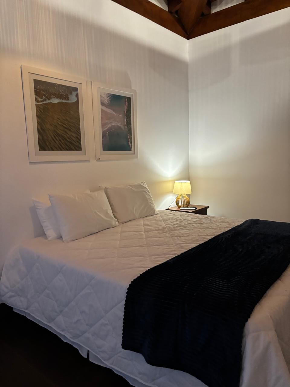 A bedroom with a bed, two framed pictures on the wall, a bedside lamp, and a dark throw blanket.