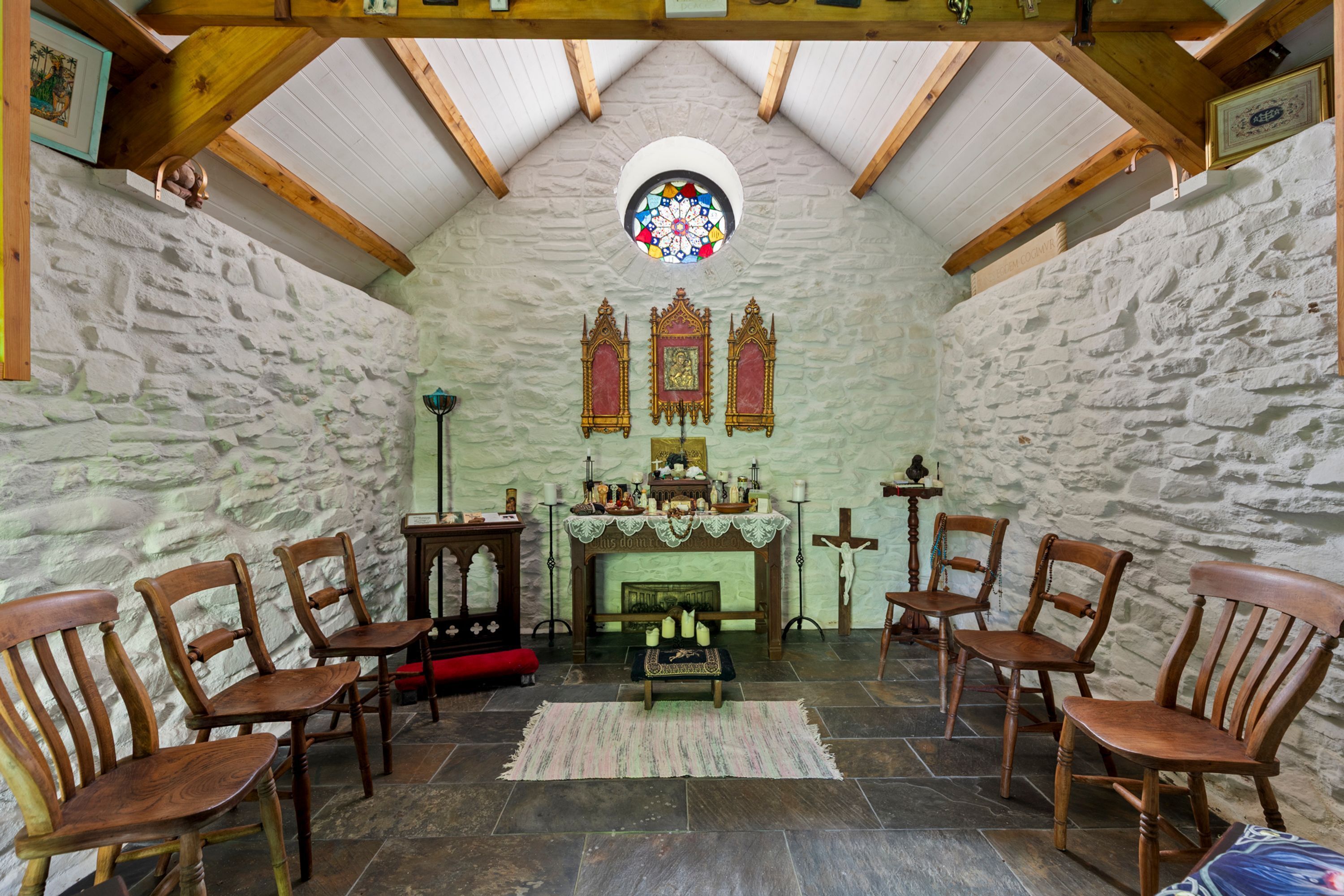 A chapel with stone walls, wooden pews, a stained glass window, and a fireplace.