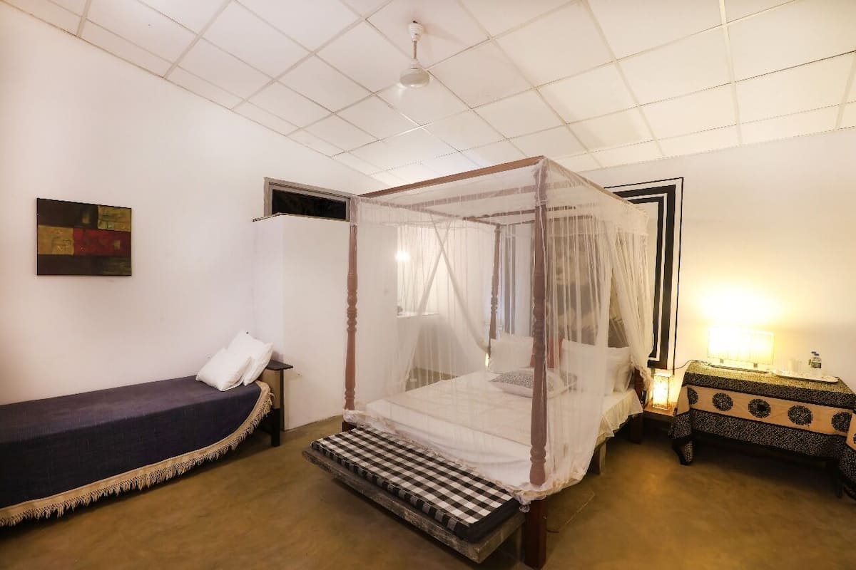 A room with a four-poster bed, a single bed, a bedside table with a lamp, and a wall-mounted painting.
