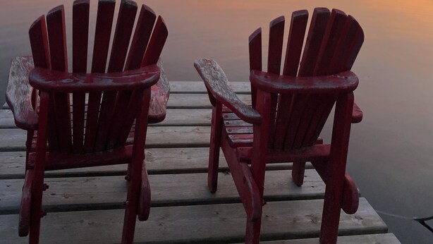 Dos sillas de madera tipo Adirondack en un muelle de madera con vista a un lago tranquilo al atardecer.