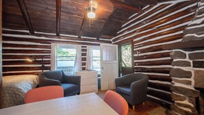 Interior de una cabaña de troncos con una mesa de comedor, dos sillones, un aparador y una ventana con cortinas.