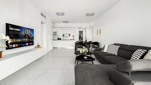 A modern living room with a flat-screen TV, a grey sofa, and a dining area with a kitchen in the background.