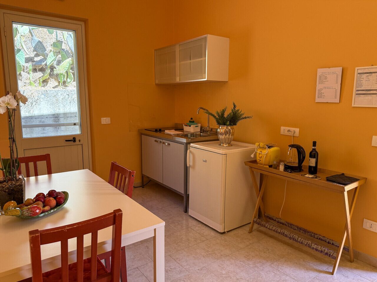 Une cuisine compacte comprenant une table à manger, des chaises, un réfrigérateur, un évier et une fenêtre donnant sur un espace verdoyant.