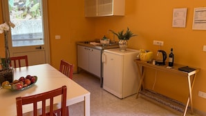 A compact kitchen with a dining table, chairs, a refrigerator, a sink, and a window with a view of greenery. - Bartoli Estate - Bouganville Apartment (Mazzarino)
