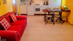Un espace de vie compact comprenant un canapé rouge, une table à manger avec quatre chaises, un coin cuisine avec des meubles blancs et un mur jaune.