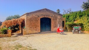 Un bâtiment rustique doté d’une grande entrée en arc de cercle, une structure au toit de chaume sur la gauche et un véhicule vert garé devant.