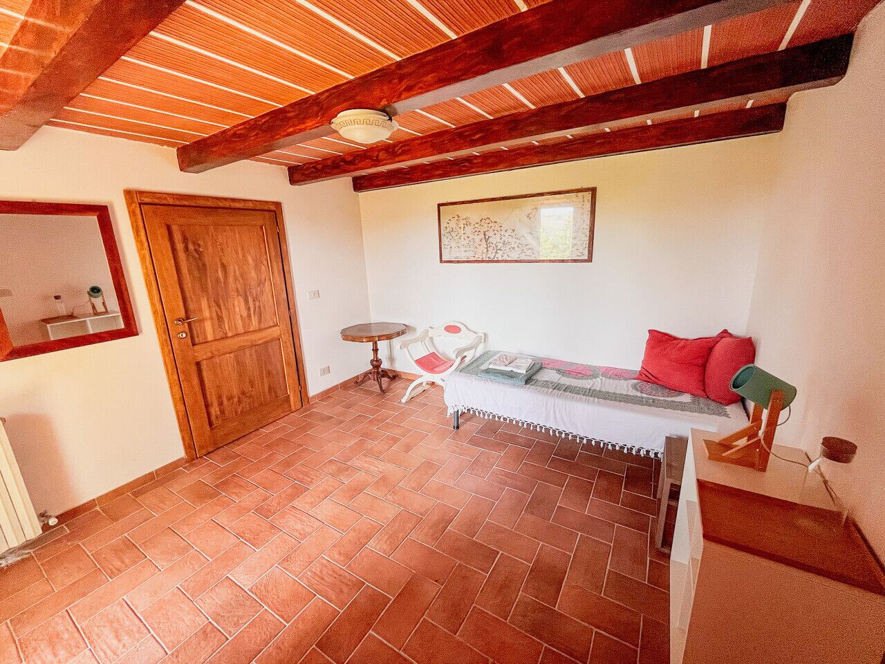 A room with a tiled floor, a wooden ceiling, a bed with pillows, a small table, and a chair.