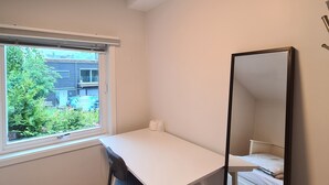 A small, bright room with a white desk, a chair, a large mirror, and a window with a view of greenery and buildings outside.