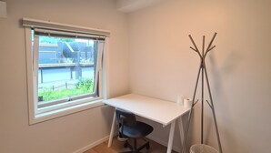 A small, well-lit room with a white desk, a black office chair, a window with blinds, a floor lamp, and a trash bin.