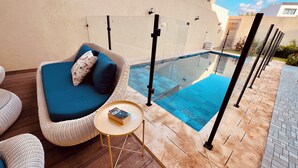 Un área de estar moderna al aire libre con un sofá y sillones de mimbre, una mesita redonda y una piscina con cubierta de vidrio.