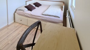Une petite chambre à coucher, bien rangée, avec un lit simple, une chaise en bois, un bureau et une fenêtre laissant entrer la lumière naturelle.