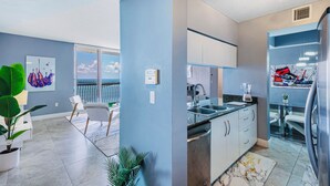 Una cocina moderna con horno empotrado, un lavamanos y un refrigerador. Hay una zona de comedor con una mesa y sillas, y un ventanal con vistas al cielo.