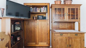 Un mueble de madera con estantes abiertos que contiene libros y objetos decorativos, un televisor montado en la pared y un banco acolchado.