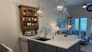Eine moderne Küche mit Kochinsel, einem Holzschrank mit Tassen, Pendelleuchten und Blick auf den Wohnbereich mit Sofa und Gemälde.