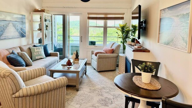 Ein geräumiges Wohnzimmer mit einer Couch, Sesseln, einem Couchtisch und einem Esstisch. Ein großes Fenster bietet Blick auf Grünflächen.