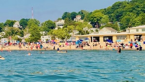 Ein voller Strand mit badenden und sonnenden Menschen, ein klarer Himmel und Gebäude im Hintergrund.
