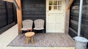 Un espacio de estar techado al aire libre con dos sillones de mimbre y una mesa redonda pequeña, una puerta blanca y un bote de basura metálico.